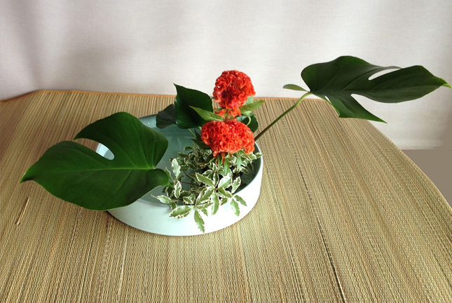 Water reflecting moribana arrangement with red flowers and monstera leaves