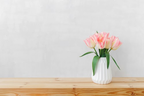 White geometric ceramic vase with bouquet of pink tulips flowers on a wooden table or shelf on a background of light gray wall.