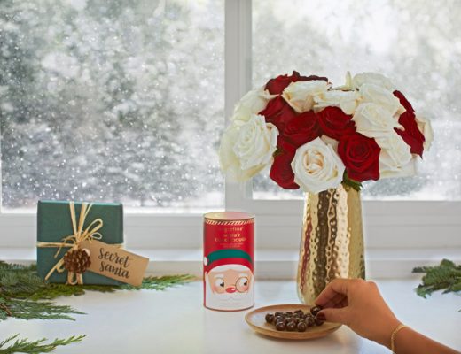 red and white flower arrangement next to a wrapped secret Santa package, greenery and Chrismas treats in front of a window.
