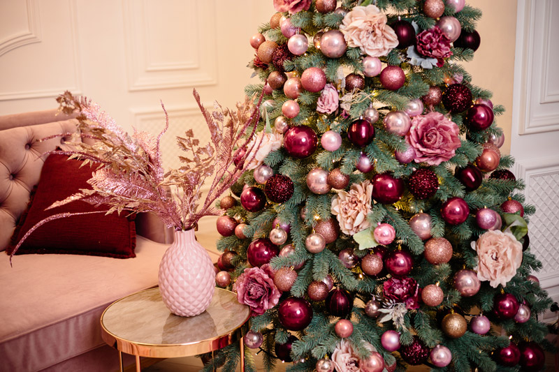 christmas tree decorated with classic sphere ornaments, tinsels, and flowers next to a vase with dried flowers on a table.